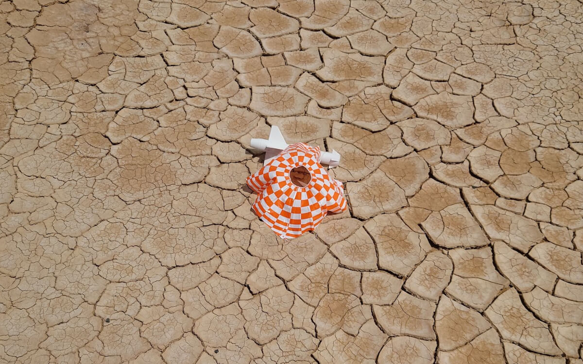 A photo of a white rocket, Frank, on a cracked, dry desert floor. The rocket's nose cone is off and its parachute has deployed. There is no damage to the rocket, but the motor is missing.